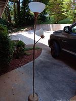 Photo of a two-arm floor lamp standing on an outdoor driveway next to a parked vehicle, showing frosted glass shades and metal stand.