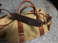 Front and angled view showing beige fabric body and brown leather handles, straps, and shoulder strap with padded section.