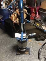 Front view of Shark upright vacuum cleaner showing whole unit, hose, and wand in storage position, with other household items in background.