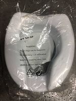 Photo of the white raised toilet seat inside sealed plastic packaging showing warning label.