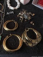Photo showing various jewelry items including gold-tone hoop earrings, a white beaded bracelet, and a cluster of small stud earrings on a black surface.