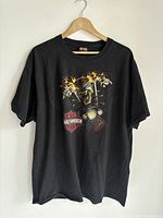 Black Harley Davidson T-shirt hung on light colored wood hanger against white wall. Front shirt graphic shows flaming skeletal rider on motorcycle handlebars with Harley-Davidson logo.