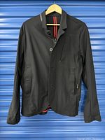 Front view of the black men's blazer showing zipper pockets and button closure, hung on a hanger against a blue backdrop.