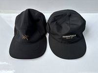 Front view of two black 5-panel hats side by side, Arc'teryx logo visible in gold on left hat, Essentials Fear of God logo printed in white on right hat.