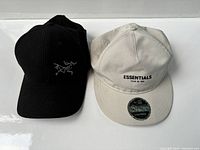 Front view of both hats showing brand logos; black Arcteryx hat and white Essentials Fear of God hat.