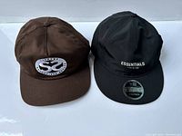 Pair of hats: brown Supreme cap and black Essentials Fear of God cap shown together.