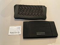 Two black clutch purses placed side by side on a flat surface, showing exterior designs. One purse has black lace over fabric, the other has a smooth, pleated design with a small metal bar.