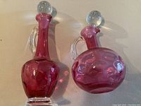 Both cranberry glass bottles shown upright with crystal lids and glass handles; surfaces glossy and in good vintage condition