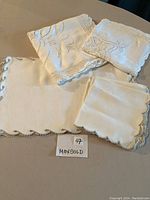 Photo showing four folded linen pieces with lace and embroidery detail, presumably tablecloths and napkins/placemats.