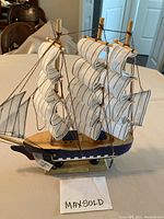 Side view of wooden model sail ship with white cloth sails and rigging on wood base, label visible