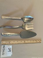 Three silver plate serving utensils laid flat on beige surface alongside ruler for scale