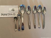 Photo showing the set of 7 small serving utensils arranged in a line with MaxSold lot number 16 card. Includes sterling silver and silver plated spoons of various shapes and decorative elements.