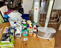 Photo showing a wooden table with multiple cleaning chemicals bottles including Bona, Scrub Free, Comet, Murphy Oil Soap, a Hoover gray upright scrubber vacuum, a white lamp with a round conical shade, and a white metal wire rack.
