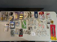 Overview of all hardware items laid out on a table including hinges, handles, hooks, catches, and miscellaneous small hardware parts in packaging and unpackaged states.