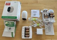Photo showing the Netgear Arlo camera, original box, instruction manuals, rechargeable batteries, chargers, USB cable and power adapter laid out on a wooden floor.
