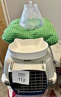 Photo showing plastic pet carrier with metal front door, green textured mat on top, ceramic cat-shaped bowls, and plastic cone collar