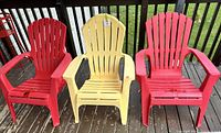Front view of three plastic Adirondack chairs: two red and one yellow, showing wear and paint marks.