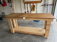 Full view of the wooden workbench showing the sturdy particle board top, thick wooden legs, and lower shelf with locking caster wheels.
