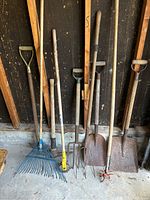View of nine garden tools including multiple shovels, pitchforks, and rakes with wooden handles and metal working parts, leaning against a wall.