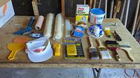 Full view of assorted handyman tools laid on wooden table, including paint rollers, brushes, putty knives, caulking gun, plaster container, adhesive box, and a white cap with Vibr logo.
