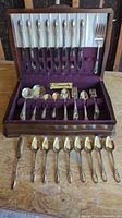 Full view of flatware set displayed in anti-tarnish lined wooden box, including knives, forks, spoons, and serving fork arranged inside and additional spoons laid out in front.