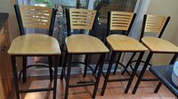 Four barstools lined up side by side on floor in front of window. Black metal frames with wood slatted backrests and wooden flat seats. Light markings on seats visible.
