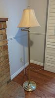 Full view of the standing brass lamp with beige lampshade showing lamp next to brick fireplace and wooden floor.
