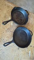 Two cast iron skillets laid on a wooden floor, showing front and back cooking surfaces and handles.