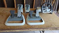 Xbox 360 racing wheel setup on a wooden surface including the wheel, two sets of pedals, and two joysticks.