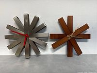 Two modernist wall clocks shown side-by-side against a white wall and gray surface. Metal clock has red hands; wooden clock has beige hands.