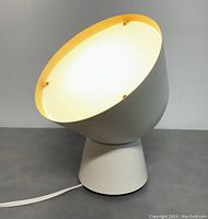 Front view of the lit lamp showing the round angled head with internal light glowing and conical base on surface.