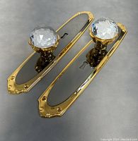 Pair of brass door handles with large faceted crystal knobs on black inset backplates with keyholes, shown from above on a grey background.