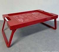 Side view of red plastic breakfast tray with folding legs extended, showing leaf pattern on tray surface and white inset handles.