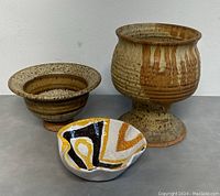 Photo showing large goblet-shaped pottery vessel, wide shallow bowl, and small abstract patterned bowl placed on grey surface against white wall.