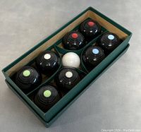 Top-down view of eight miniature black carpet bowls with colored centers (red, blue, white, green) and one white jack ball arranged inside original green box with compartment dividers.