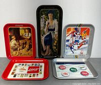Front view of 5 Coca-Cola collectible trays in various sizes and designs on gray surface