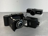 Photo of Keystone Zoom 66 camera, Kodak folding film camera and flash unit arranged on a clear stand.