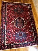 Full view of a 100% wool Persian rug showing red base with blue and cream geometric and floral patterns and fringed edges.