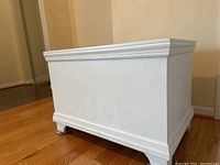 Side angle view showing the white painted wooden storage chest with decorative molding and carved feet.