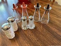 Photo of all eight salt and pepper shakers on wooden table, showing both milk glass Fenton shakers and clear glass Pyrex shakers with chrome and black tops.
