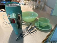 Side view of GE mint green hand mixer with two metal beaters sitting on a counter, accompanied by Fire King mint green teacup, saucer and bowl in the background.