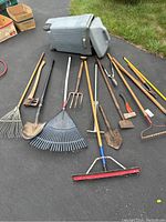 Full view of all garden and lawn tools laid on driveway near grass with rolling trash bin tipped over