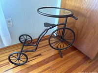 Full view of a metal bicycle plant stand with round glass tabletop placed on wooden floor against wall and door.