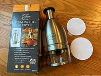 Photo of the Crofton Stainless Steel Food Chopper alongside its original box and two round rubber covers on a wooden surface.