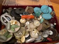 Close-up of mixed assorted buttons in a clear plastic box, showing variety of sizes, colors, and materials including fabric-covered turquoise buttons.