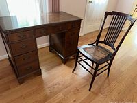 Full view of dark wood kneehole desk alongside matching wooden chair with cane seat on hardwood floor.