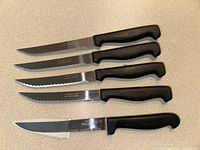 Four Tramontina steak knives with serrated blades and black handles, plus one EKCO Waverex serrated cutting knife, displayed on countertop.