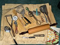 Top-down view of collection of vintage kitchen utensils on beige pastry cloth including wooden rolling pin, metal ladles, potato masher, whisks, peelers, and other hand tools.