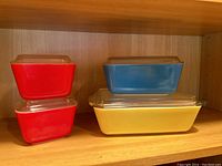 Stack of two red Pyrex dishes with glass lids, next to stack of blue on yellow dish with lids on a wooden shelf