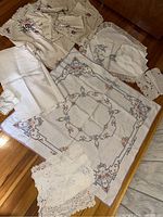 Overview photo of all linens arranged on wooden floor. Includes large embroidered tablecloth and smaller tatted and lace-edged linens.